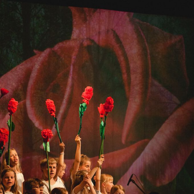 Mitglieder Des Kinderchores Halten Selbstgebastelte Rosen In Den Händen, Im Hintergrund Die Projektion Einer Großen Rosenblüte