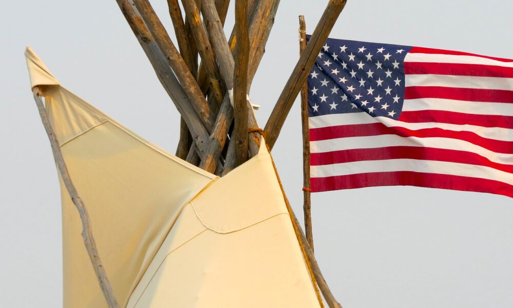 Spitze eines Indianerzelts mit US-Flagge im Hintergrund